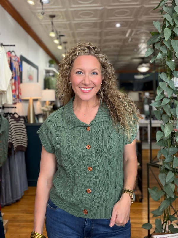 A smiling woman with curly blonde hair wearing a sage green cable-knit short-sleeve button-up cardigan and blue jeans, standing inside a retail store.