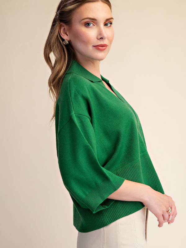 A woman wearing a bright green knit polo-style sweater with three-quarter sleeves and a relaxed fit, paired with off-white pants, photographed against a plain beige studio background.