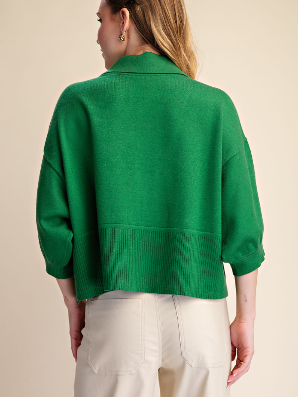 A woman shown from the back wearing a bright green knit cropped sweater with three-quarter sleeves and ribbed trim, paired with light beige pants, against a solid beige background.