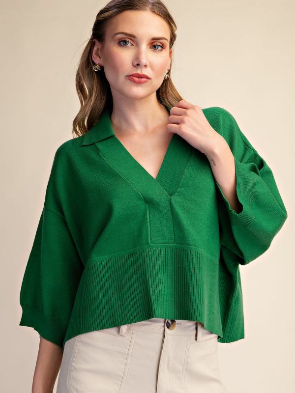 A woman wearing a vibrant green knit polo sweater with a V-neck and wide three-quarter sleeves, paired with light beige trousers, set against a plain beige studio background.