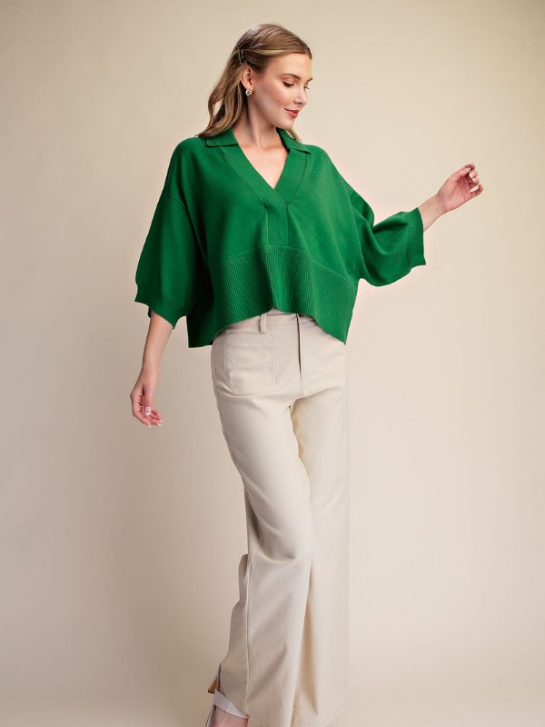 A woman modeling a vibrant green, oversized short-sleeve blouse with a V-neck and wide sleeves, paired with cream-colored wide-leg trousers, against a solid beige studio background.