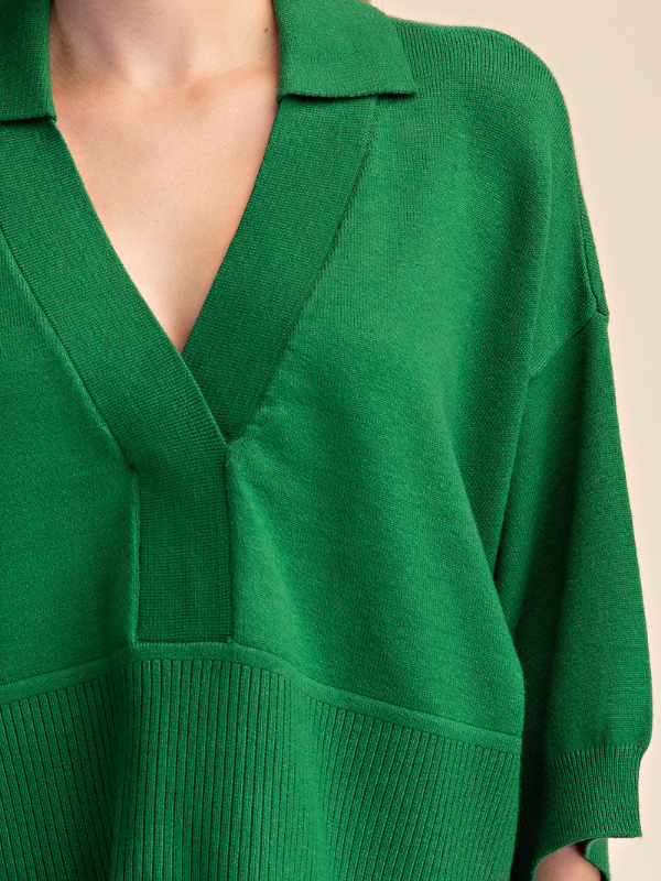 Close-up of a person wearing a bright green knit sweater with a V-neck and short sleeves, against a plain light beige background.