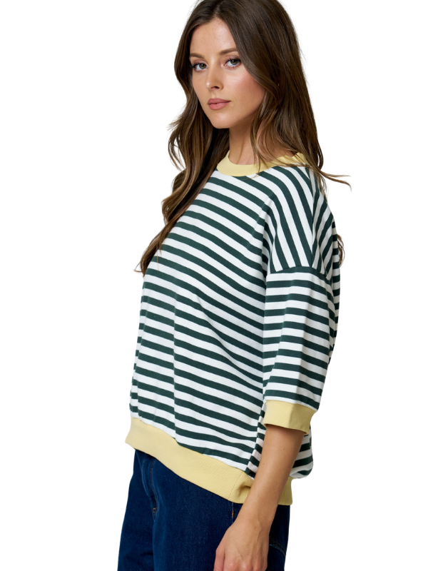 A woman modeling a loose-fitting knit top with horizontal dark green and white stripes, featuring solid pale yellow trim at the crew neck, cuffs, and hem, paired with dark blue jeans against a plain white background.