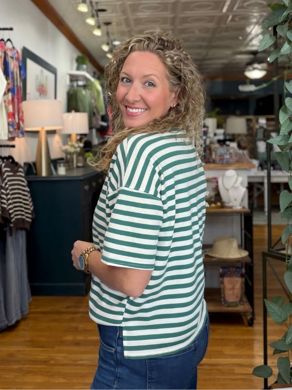 A woman with curly blonde hair smiling and looking back over her shoulder while wearing a green and white horizontally striped short-sleeve t-shirt and blue jeans inside a retail store.