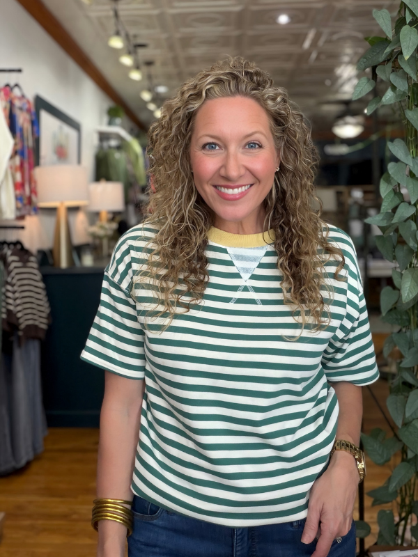 A smiling woman with curly blonde hair wearing a green and white striped t-shirt with a yellow collar, standing inside a boutique.