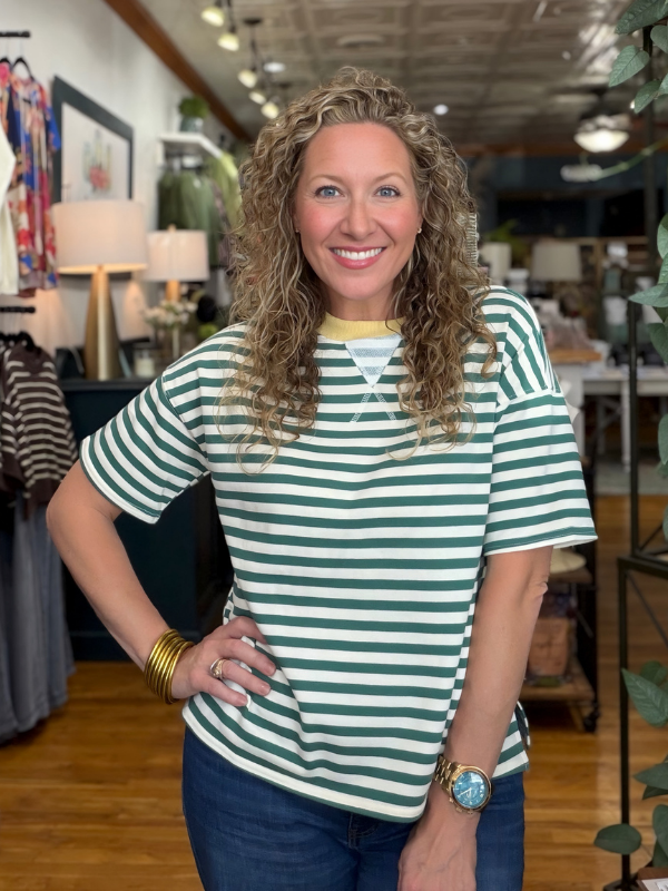 A smiling woman with curly blonde hair stands in a boutique wearing a green and white striped short-sleeve t-shirt with a yellow inner collar, paired with dark blue jeans.