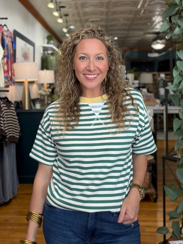 A smiling woman with curly blonde hair stands in a boutique wearing a green and white horizontally striped short-sleeve t-shirt with a yellow collar trim and blue jeans.