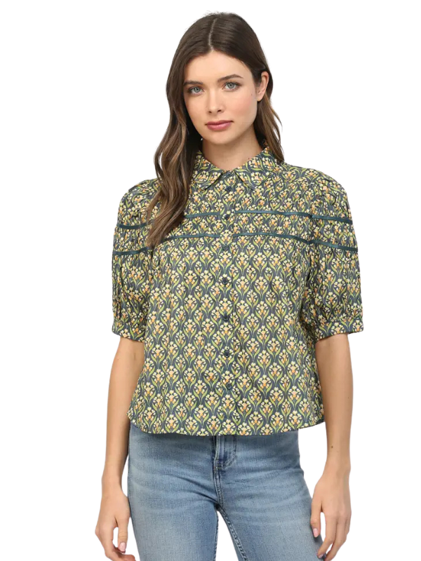 A woman modeling a short-sleeve button-up blouse with a green, yellow, and white geometric pattern, paired with blue jeans, against a plain white background.