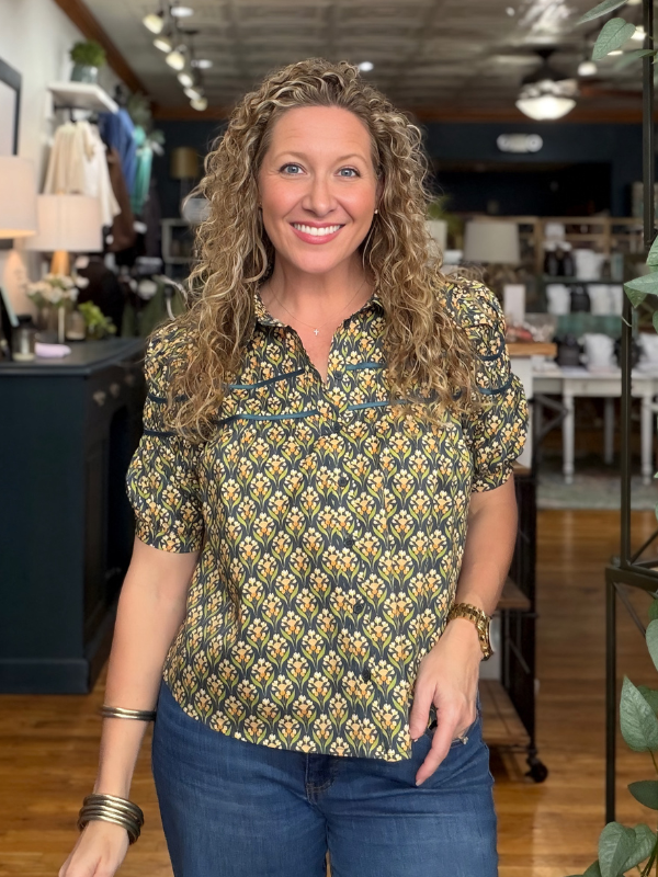 A smiling woman with curly blonde hair stands in a boutique wearing a short-sleeve button-up shirt with a yellow, green, and black geometric pattern, paired with blue jeans.