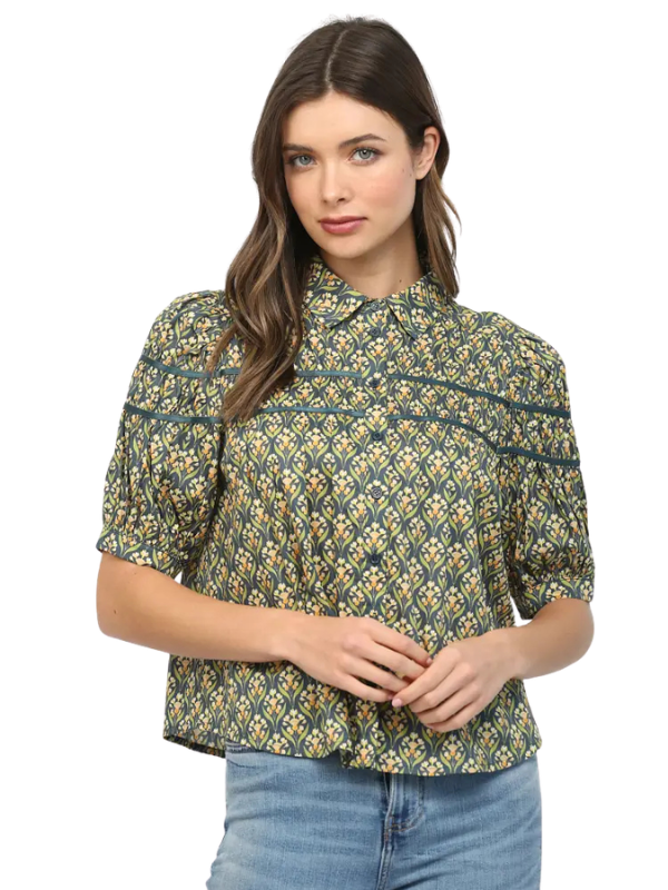A woman modeling a short-sleeve button-up blouse with a green, yellow, and white geometric pattern, paired with blue jeans against a plain white background.
