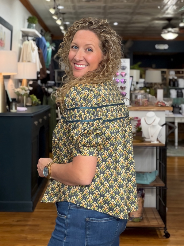A woman with curly blonde hair wearing a green and yellow patterned short-sleeve blouse with blue jeans, standing in a retail store.