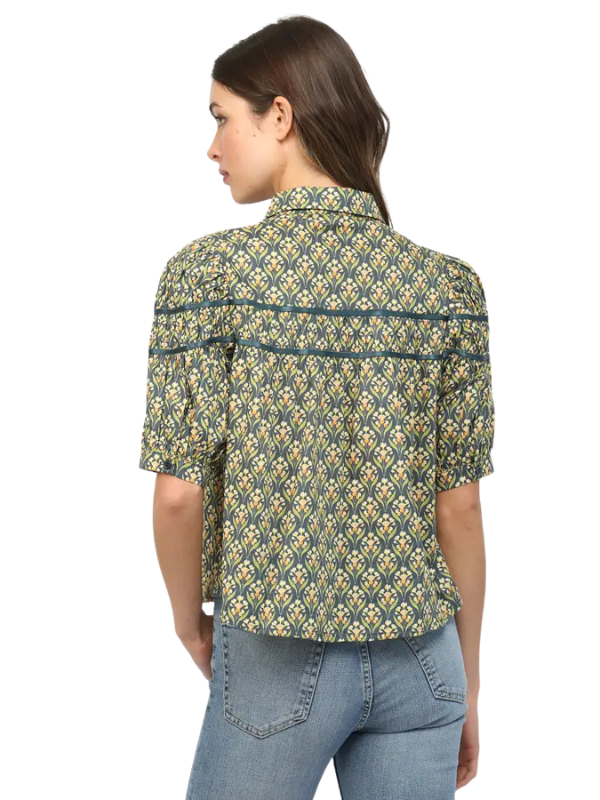 A woman shown from the back wearing a short-sleeve button-up shirt with a green, yellow, and blue geometric pattern, paired with light blue jeans, against a plain white background.