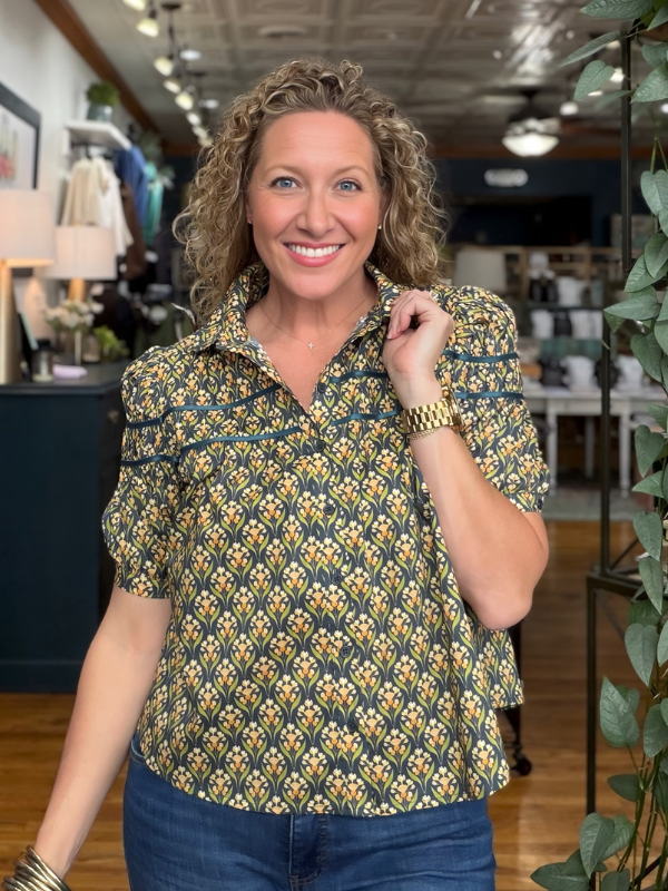 A smiling woman with curly blonde hair stands in a retail store wearing a short-sleeve collared blouse with a yellow and green floral pattern, paired with blue jeans.