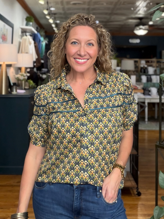 A smiling woman with curly blonde hair wearing a short-sleeve button-up shirt with a yellow, green, and blue geometric pattern, paired with blue jeans, standing inside a retail store.