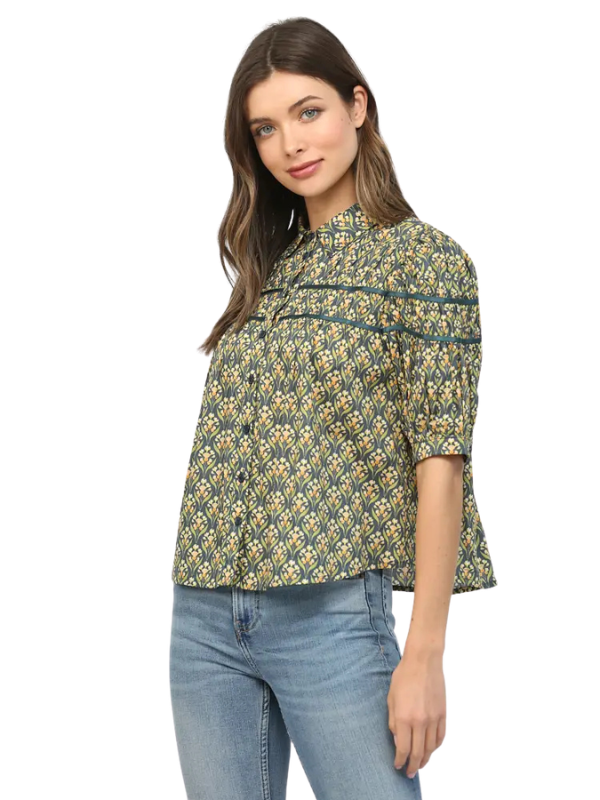 A woman modeling a short-sleeve button-up blouse with a green, yellow, and white geometric pattern, paired with blue jeans, against a plain white background.