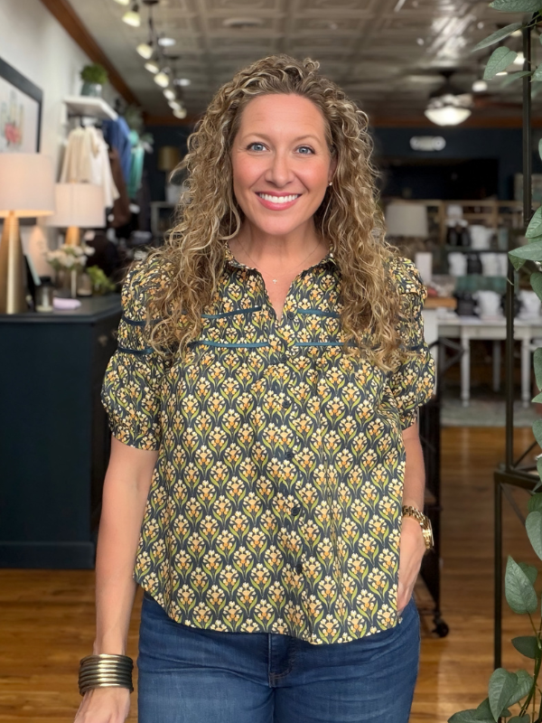 A smiling woman with curly blonde hair stands in a boutique wearing a short-sleeve button-up blouse with a yellow and green geometric pattern, paired with blue jeans.