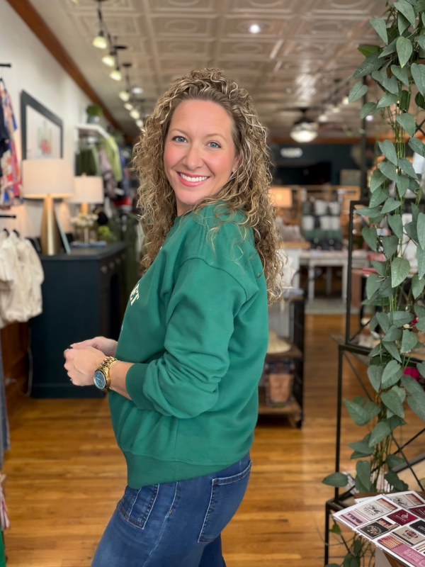A smiling woman with curly blonde hair wearing a green crewneck sweatshirt and blue jeans, standing in a retail store interior with wood floors and shelves in the background.