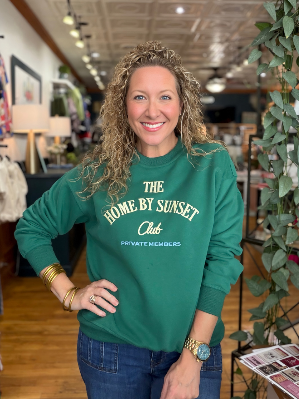 A smiling woman with curly blonde hair stands in a retail store wearing a green crewneck sweatshirt with cream text that reads 'THE HOME BY SUNSET Club PRIVATE MEMBERS'.
