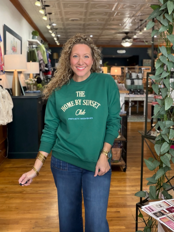 A woman with curly hair stands in a retail store wearing a green crewneck sweatshirt with the text 'THE HOME BY SUNSET Club' printed in white on the front, paired with blue jeans.