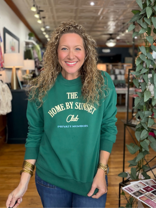 A smiling woman with curly blonde hair wearing a green crewneck sweatshirt with cream text that reads 'THE HOME BY SUNSET Club PRIVATE MEMBERS' and blue jeans, standing inside a retail store.