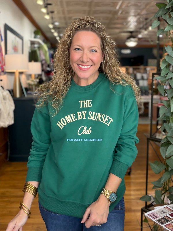 A smiling woman with curly blonde hair stands in a retail store wearing a green crewneck sweatshirt with cream text that reads 'THE HOME BY SUNSET Club PRIVATE MEMBERS'.