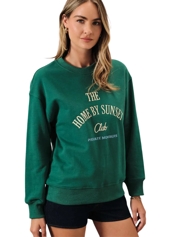 A woman wearing a dark green crewneck sweatshirt with cream-colored text that reads 'THE HOME BY SUNSET Club' and 'PRIVATE MEMBERS' on the front, paired with black shorts, against a plain white background.