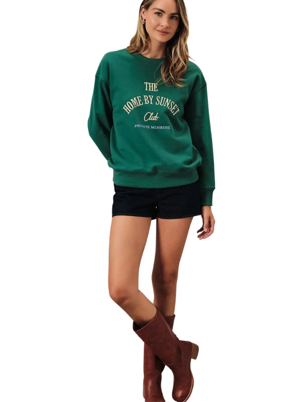 A woman modeling a dark green crewneck sweatshirt with cream-colored text reading 'THE HOME BY SUNSET Club' and 'Pittsford Michigan' on the front, paired with black shorts and brown boots against a plain white background.