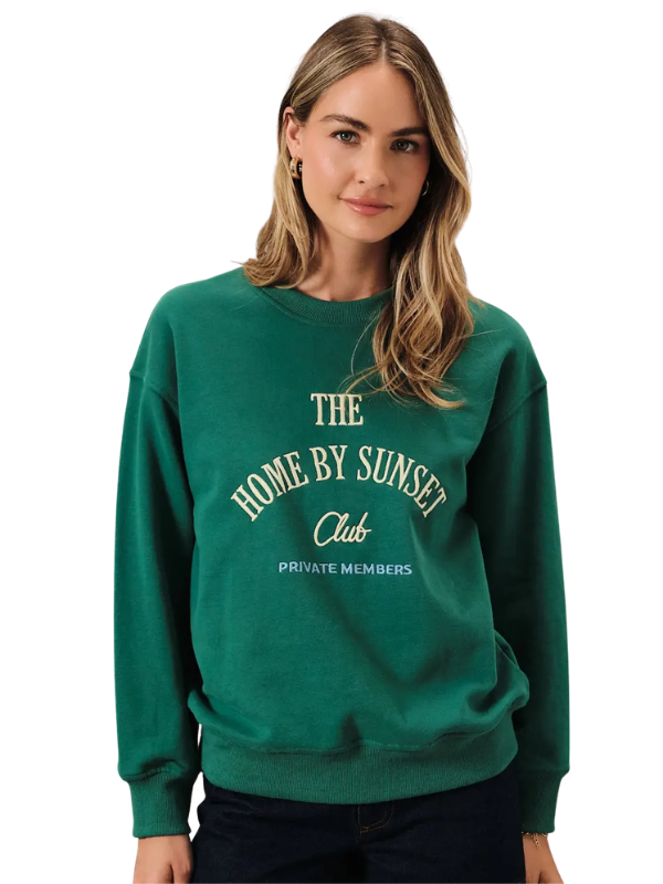 A woman wearing a dark green crewneck sweatshirt with cream-colored text that reads 'THE HOME BY SUNSET Club PRIVATE MEMBERS' on the front, set against a plain white background.