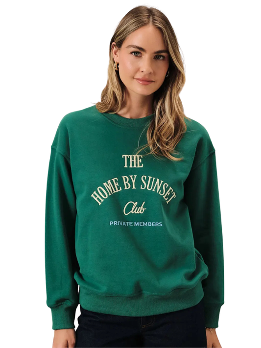 A woman wearing a dark green crewneck sweatshirt with cream-colored text that reads 'THE HOME BY SUNSET Club PRIVATE MEMBERS' on the front, set against a plain white background.