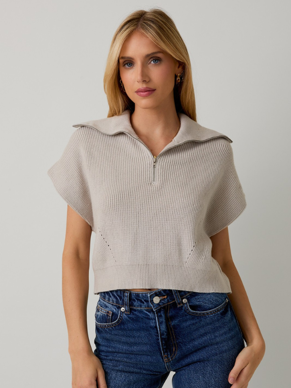 Woman wearing a beige short-sleeve sweater and blue jeans against a plain background