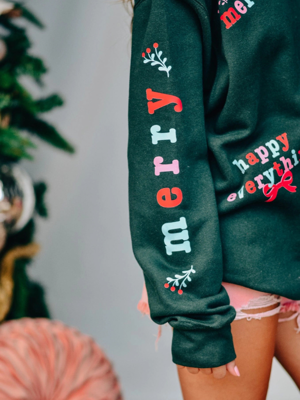 Green sweatshirt with festive text and designs worn by a person, Christmas tree in the background