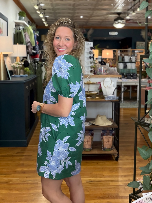 A woman with curly blonde hair wearing a green short-sleeve dress with a light blue floral pattern, standing inside a retail store.
