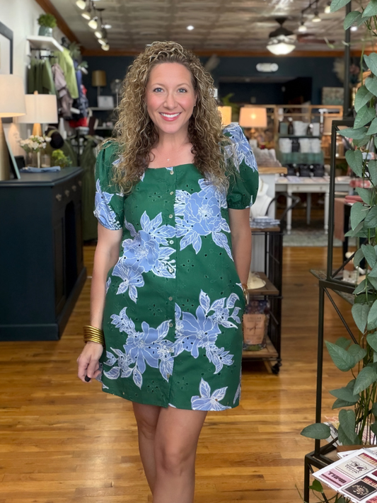 A woman with curly blonde hair wearing a green and light blue floral puff-sleeve dress, standing in a retail store with wooden floors and home decor items in the background.