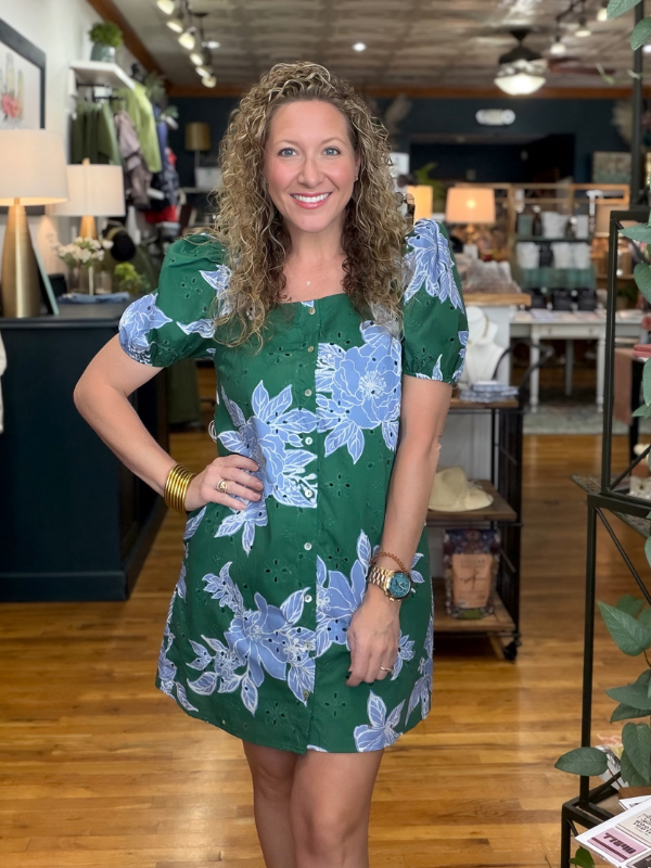 A woman with curly brown hair stands in a boutique wearing a green short-sleeve dress with a light blue floral print. The dress features eyelet eyelet fabric and puff sleeves.