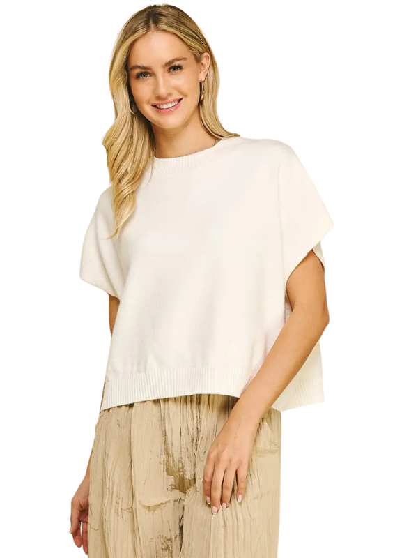 A woman modeling a cream-colored short-sleeve knit top with a relaxed fit, paired with pleated beige trousers, against a plain white background.
