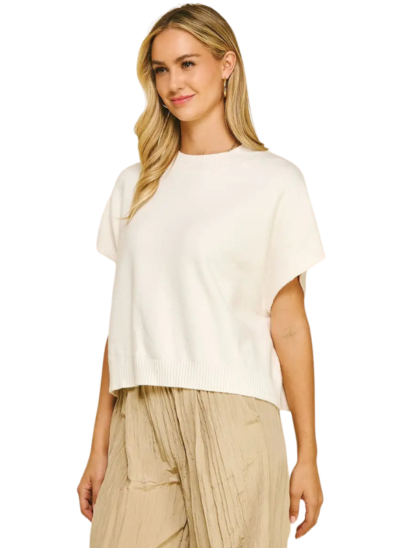A woman wearing a cream-colored short-sleeve knit top and beige pleated pants, photographed against a plain white background.