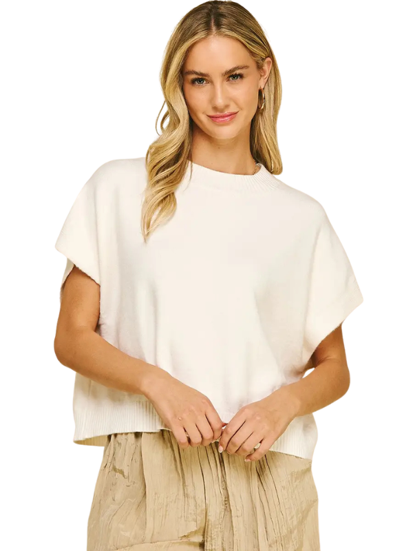 A blonde woman wearing a cream-colored short-sleeve knit top and beige pleated pants, standing against a plain white background.