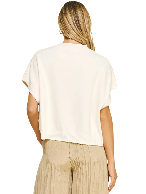A woman shown from the back wearing a cream-colored short-sleeve knit top and beige pleated trousers against a plain white background.