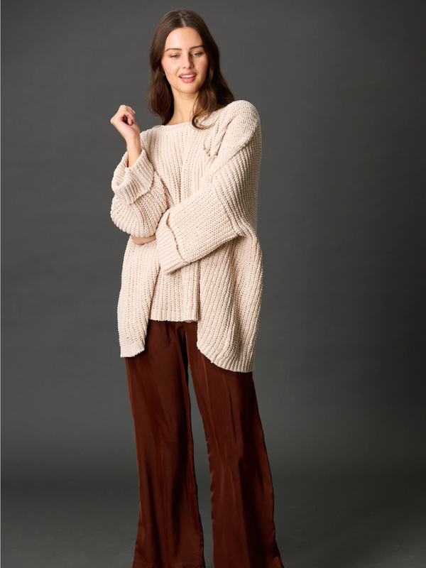 Woman wearing a beige sweater and brown pants against a dark background