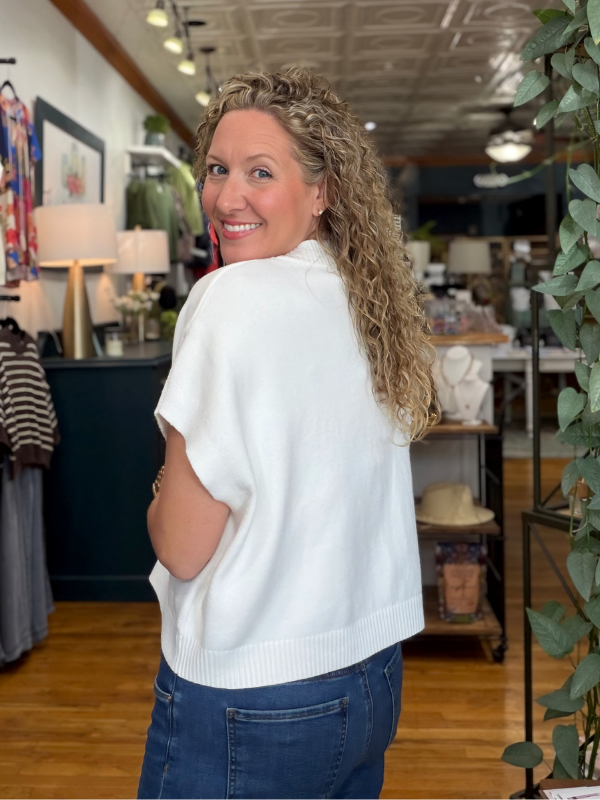 A woman with curly blonde hair wearing a white short-sleeve knit top and blue jeans, photographed from the back inside a retail store.