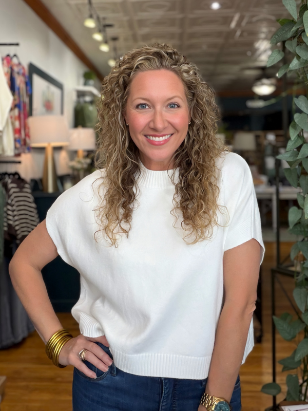 A smiling woman with curly blonde hair wearing a white short-sleeve knit sweater and blue jeans, standing in a retail store interior.