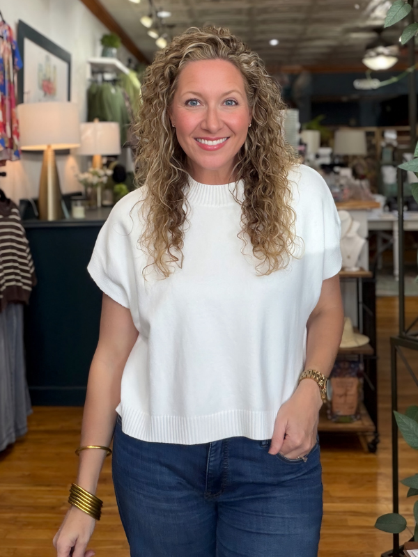 A smiling woman with curly blonde hair wearing a white short-sleeve knit top and dark blue jeans, standing in a retail store interior.