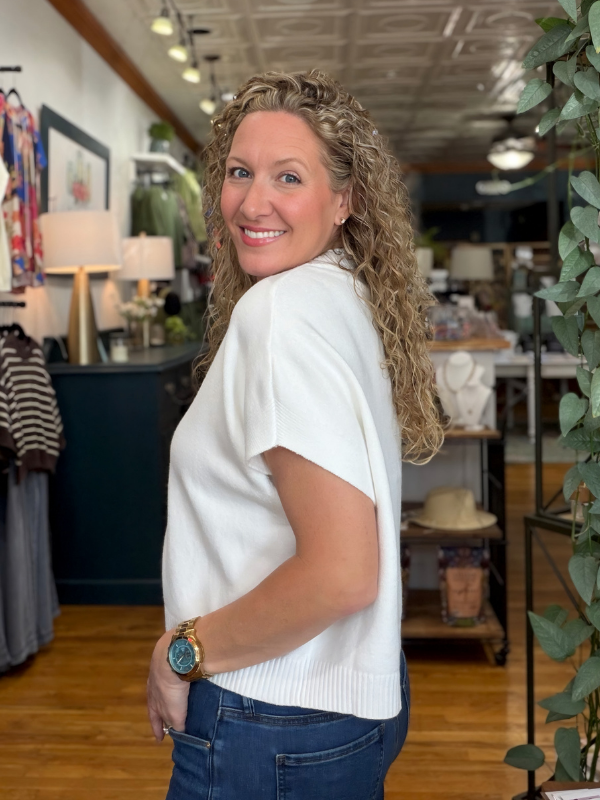 A woman with curly blonde hair wearing a white short-sleeve knit top and blue jeans, standing inside a retail store.