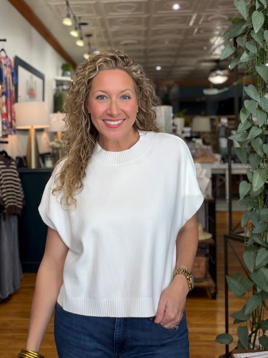 A smiling woman with curly blonde hair wearing a white short-sleeve knit top and dark blue jeans, standing inside a retail store with plants and furniture in the background.