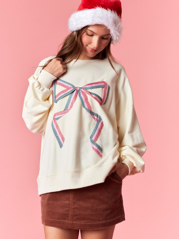 Person wearing a cream sweatshirt with a large bow design and a Santa hat on a pink background
