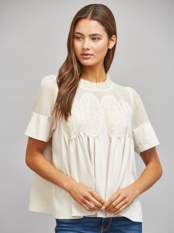 A woman modeling a short-sleeve cream blouse with sheer mesh sleeves and a floral lace applique across the chest, paired with blue jeans against a solid gray studio background.