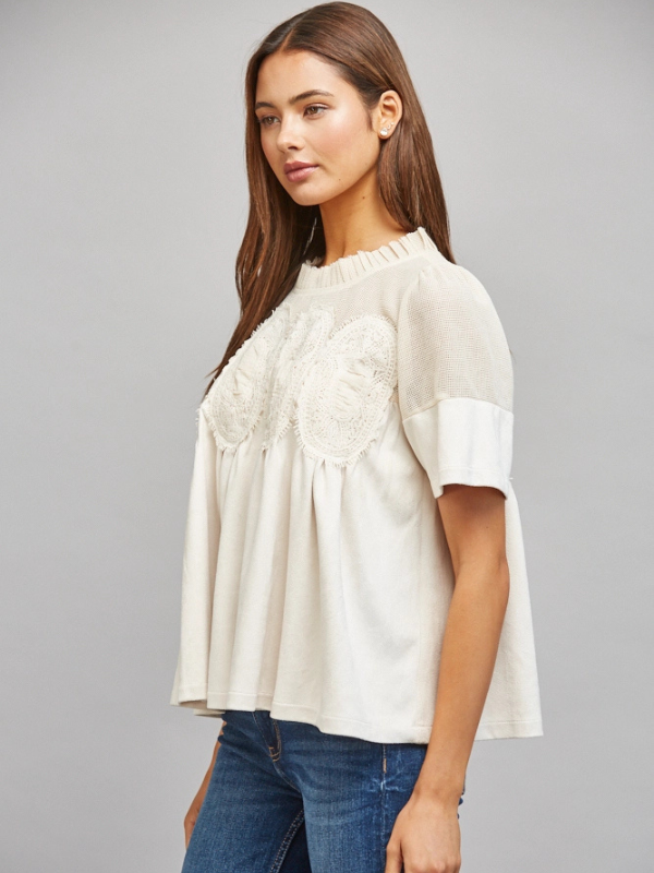 A woman modeling a short-sleeve cream blouse with delicate lace applique across the chest and ruffled detailing at the neckline, paired with blue jeans against a solid gray studio background.