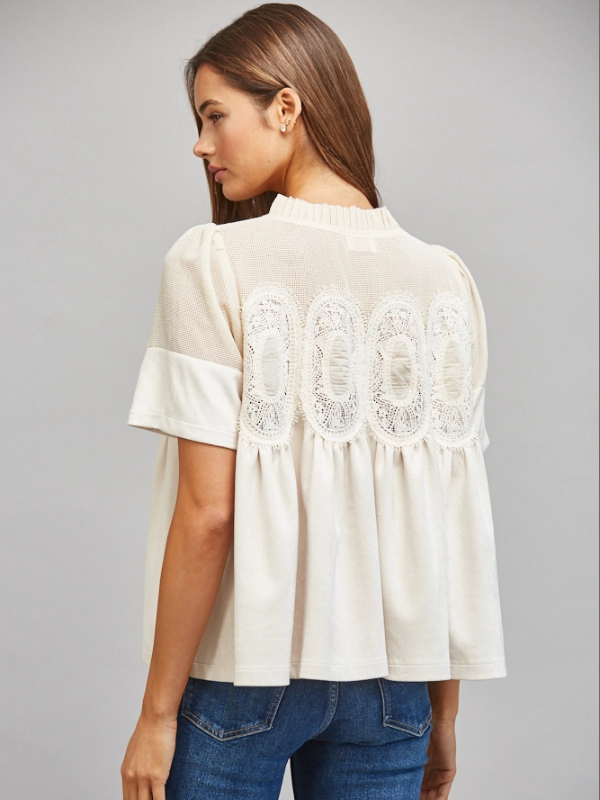 A woman shown from the back wearing a cream-colored short-sleeve blouse with lace applique across the upper back and blue jeans, set against a solid gray studio background.