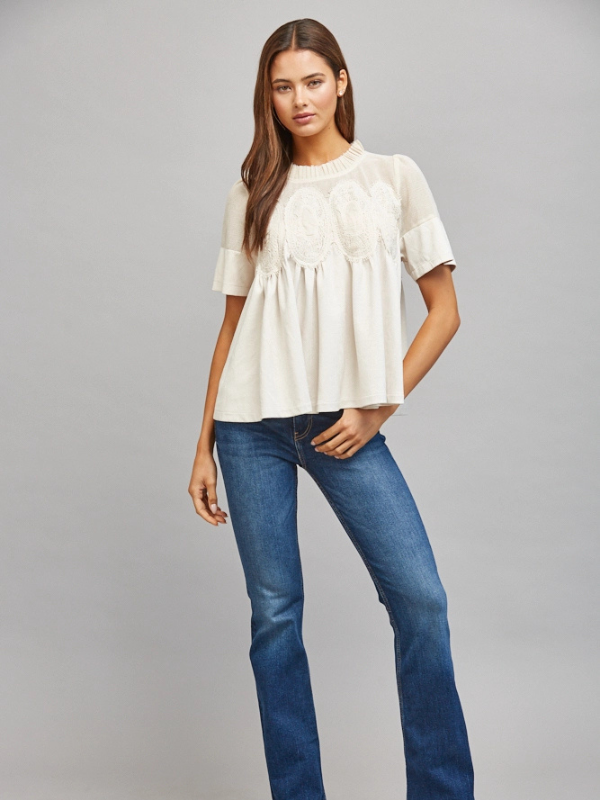 A woman modeling a short-sleeve cream blouse with delicate lace embroidery on the bodice and ruffled sleeves, paired with blue jeans, against a solid gray studio background.