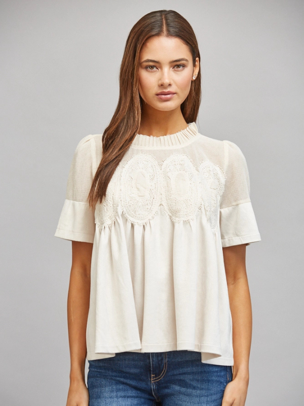 A woman modeling a short-sleeve cream blouse with delicate lace applique across the chest and ruffled detailing at the neckline, paired with blue jeans against a solid gray studio background.